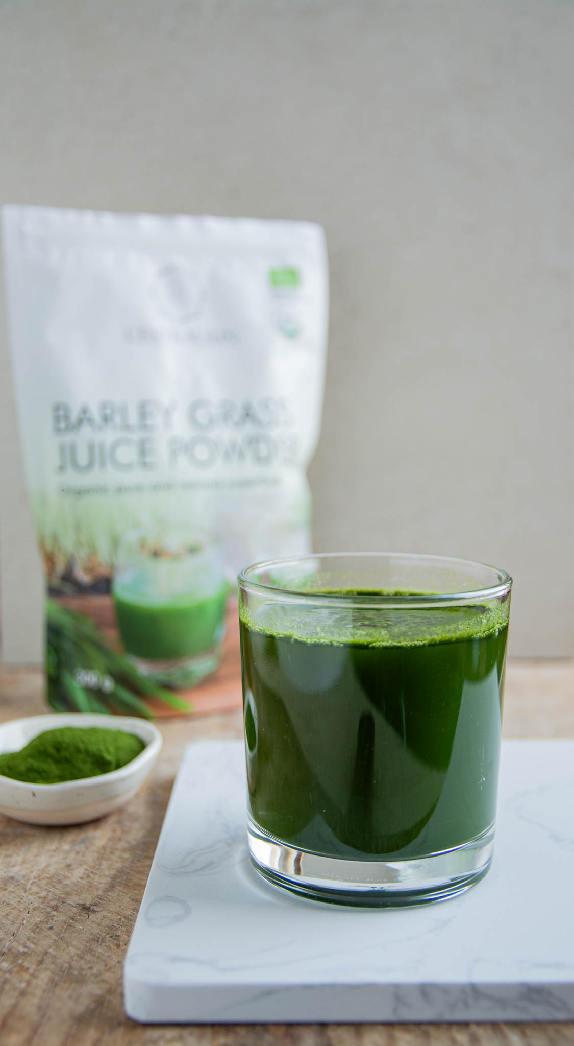 A glass of freshly mixed barley grass juice on a marble coaster, with a bowl of barley grass powder and a Centralsun barley grass juice powder package in the background.